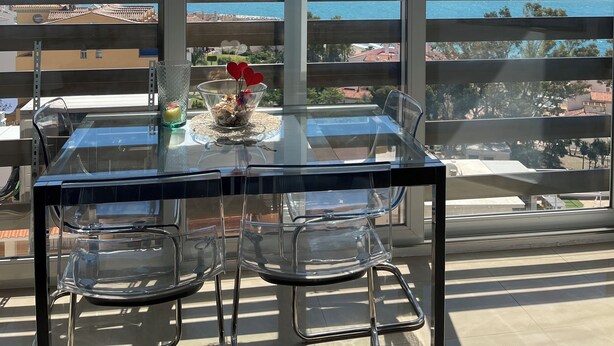 Un balcon avec une table et des chaises en verre, offrant une vue sur la côte, avec des bâtiments et un pont.