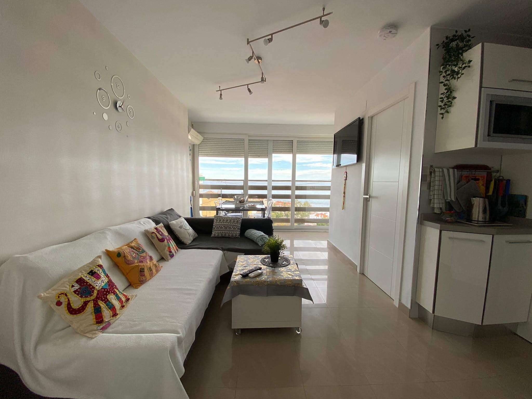 A modern living room with a sofa, a coffee table, and a kitchen area with cabinets and a TV.