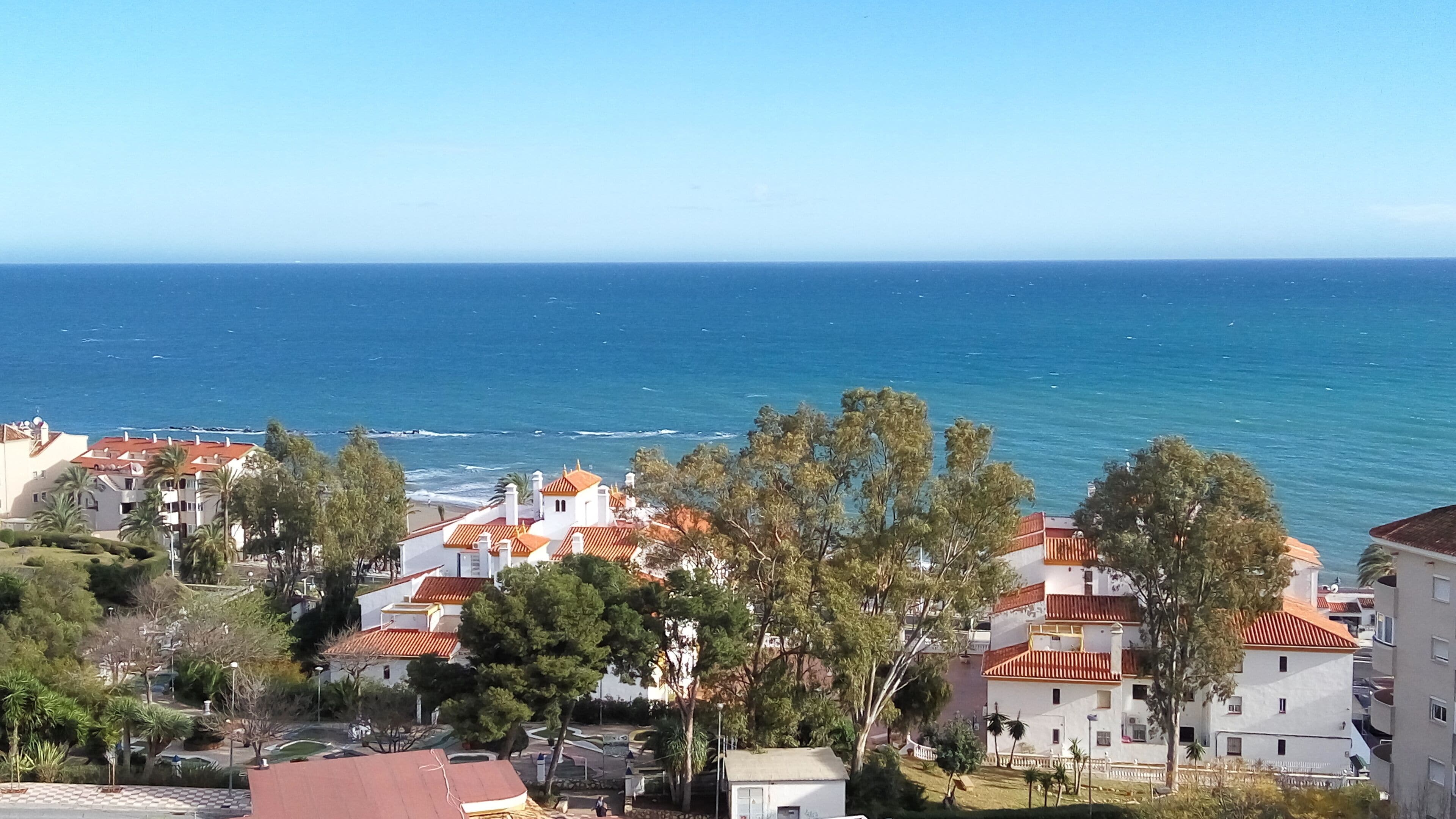 A coastal residential area with buildings featuring red-tiled roofs, surrounded by greenery and overlooking the sea.