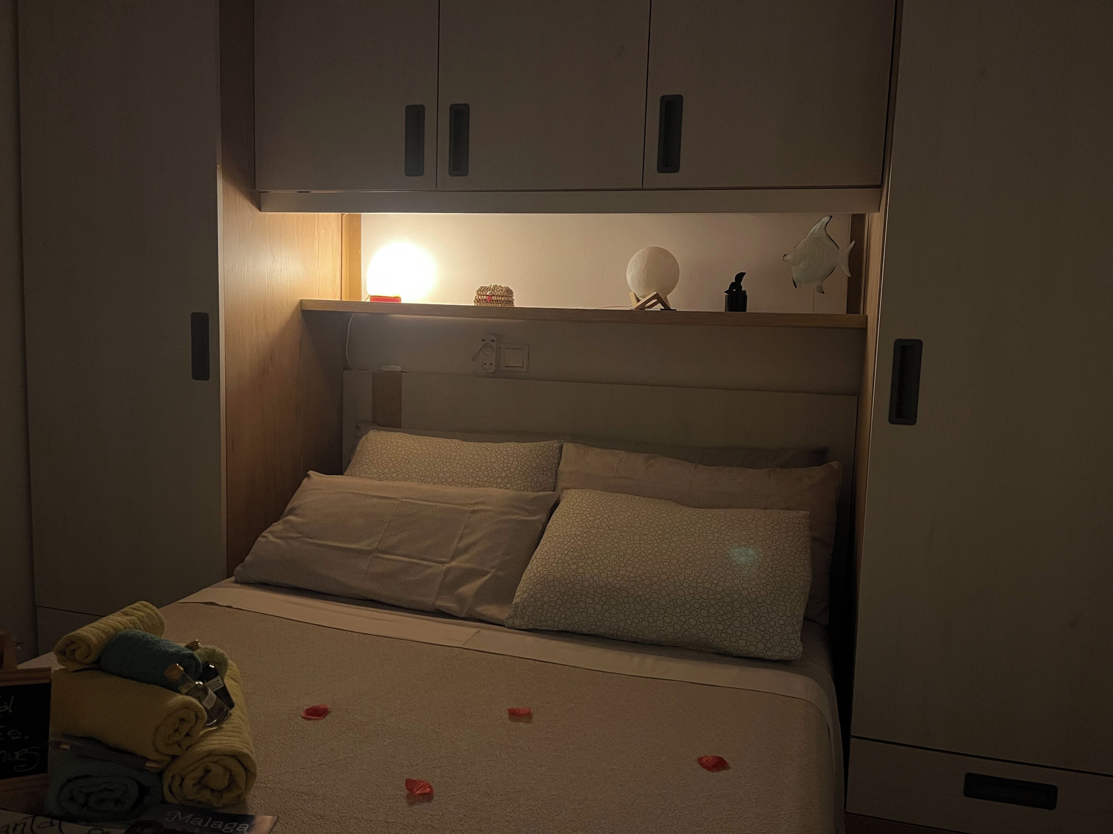 A single bed with two pillows, a bedside table with a lamp, and a shelf above the bed.