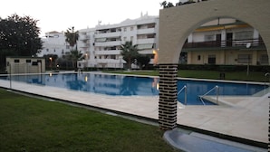 Une piscine à l’eau d’un bleu limpide, entourée d’une allée pavée. Les bâtiments adjacents sont dotés de nombreux balcons, et un petit jardin, avec de la pelouse et quelques arbres, complète l’ensemble.