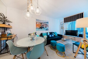 A modern living room with a round glass table, blue chairs, a dark sofa, and a TV. - 2 Room Apartment - 5 minutes to Costa da Caparica Beach and Bars (Costa da Caparica)
