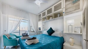 Un dormitorio con colcha y sillón color turquesa, y un aparador blanco con un florero.
