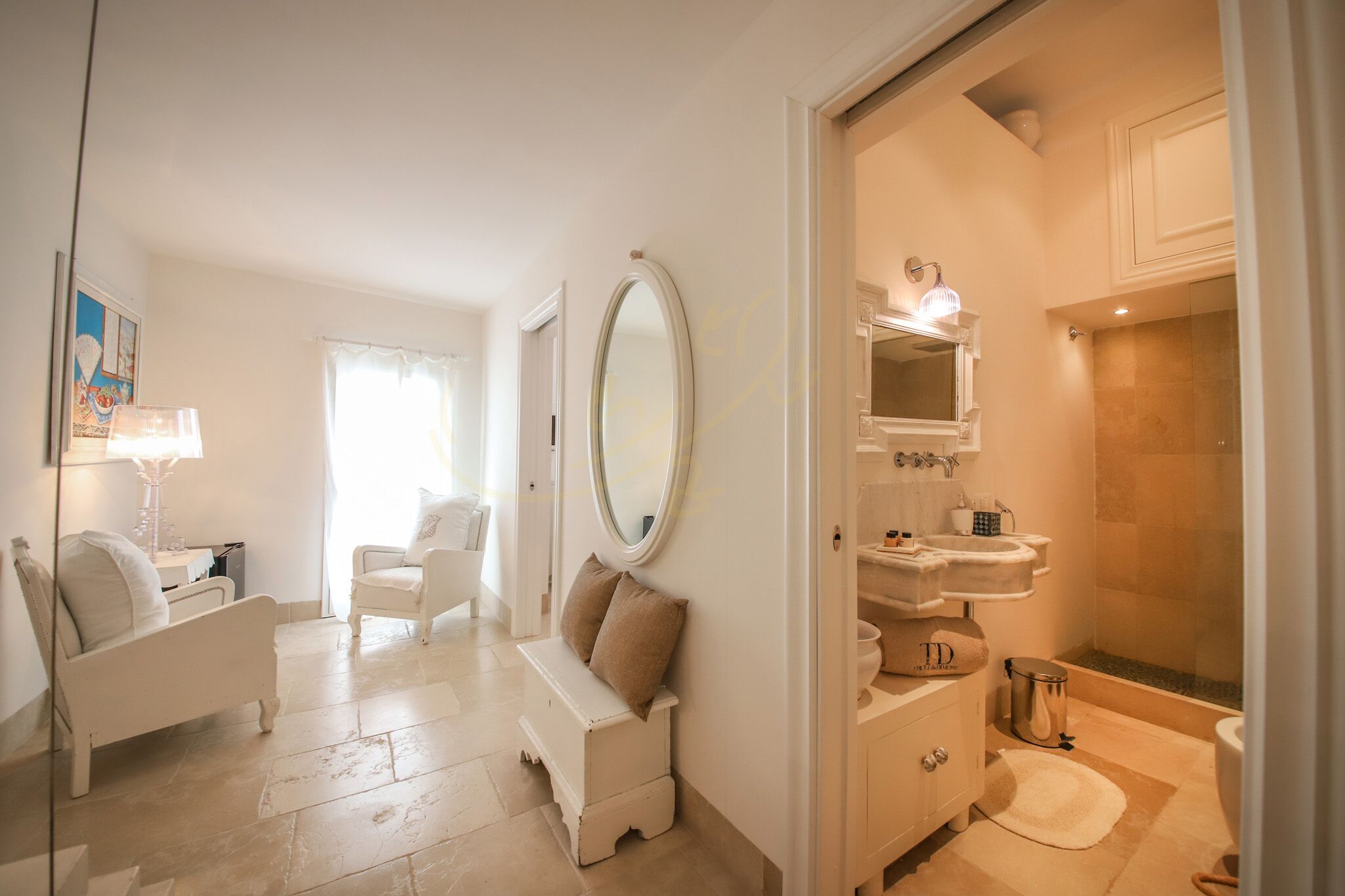 A hotel room with a large mirror, a white bench, a bedside table, a lamp, a sofa, and a bathroom with a bathtub and sink.