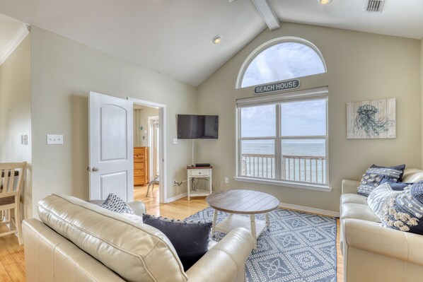 A spacious living room with a large window overlooking the beach, a comfortable seating area, and a flat-screen TV mounted on the wall. - Beautifully Decorated, Comfortable, Beachfront 3 Bedroom House on Gulf Beach (Dauphin Island)