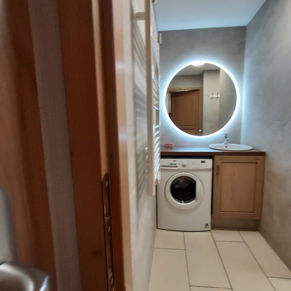 Un baño pequeño con lavadora, lavamanos y espejo con luz circular.