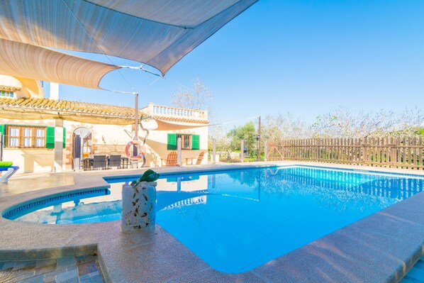 A swimming pool with a sunshade, outdoor seating, and a building with green shutters.