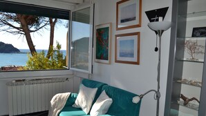 Een smaragdgroene bank met witte kussens, een vloerlamp en een raam met uitzicht op zee en bomen.