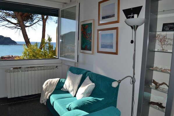 A teal sofa with white cushions, a floor lamp, and a window with a view of the sea and trees. - Quiet apartment surrounded by the green and very close to the sea (Sestri Levante)