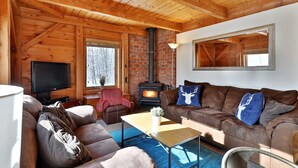 Un accogliente interno di una baita con soffitto in legno, camino, televisore e divano con cuscini decorativi.