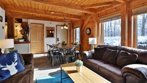 Un accogliente interno di una baita con soffitto in legno, una zona pranzo con tavolo e sedie in legno, un divano in pelle marrone e un'ampia finestra con vista innevata.