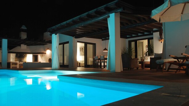 Ein Schwimmbecken bei Nacht mit blau beleuchtetem Rand, eine überdachte Terrasse mit Sitzgelegenheiten und ein gut beleuchteter Innenbereich.
