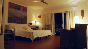 Ein Hotelzimmer mit Bett, Sessel, Tisch, Lampe, einem Gemälde und einem Deckenventilator.