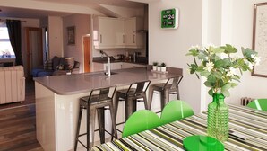 Cozinha moderna com armários brancos, uma ilha central com banquetas, um relógio verde e um vaso de planta.