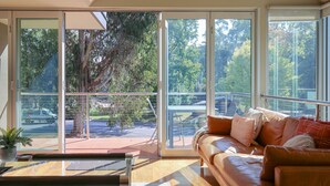 A modern living room with a leather sofa, a glass door leading to an outdoor balcony, and a view of trees and a parked vehicle. - THE GLASS PINNACLE : Bright (Bright)