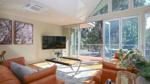 A spacious living room with a large TV, comfortable seating, and a balcony with a view of trees and a road. - THE GLASS PINNACLE : Bright (Bright)