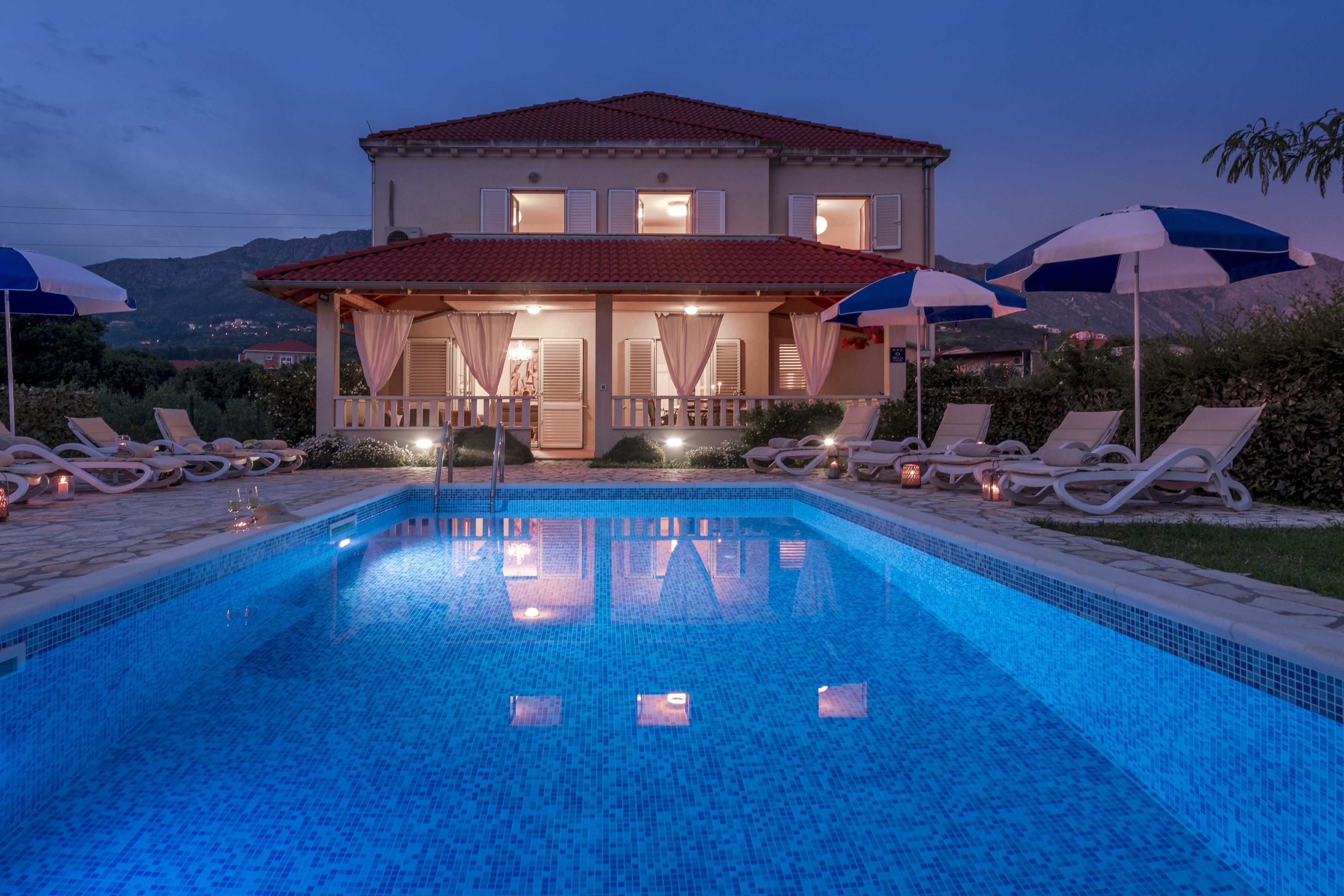 Uma piscina com espreguiçadeiras e guarda-sóis, uma casa com varanda e montanhas ao fundo.