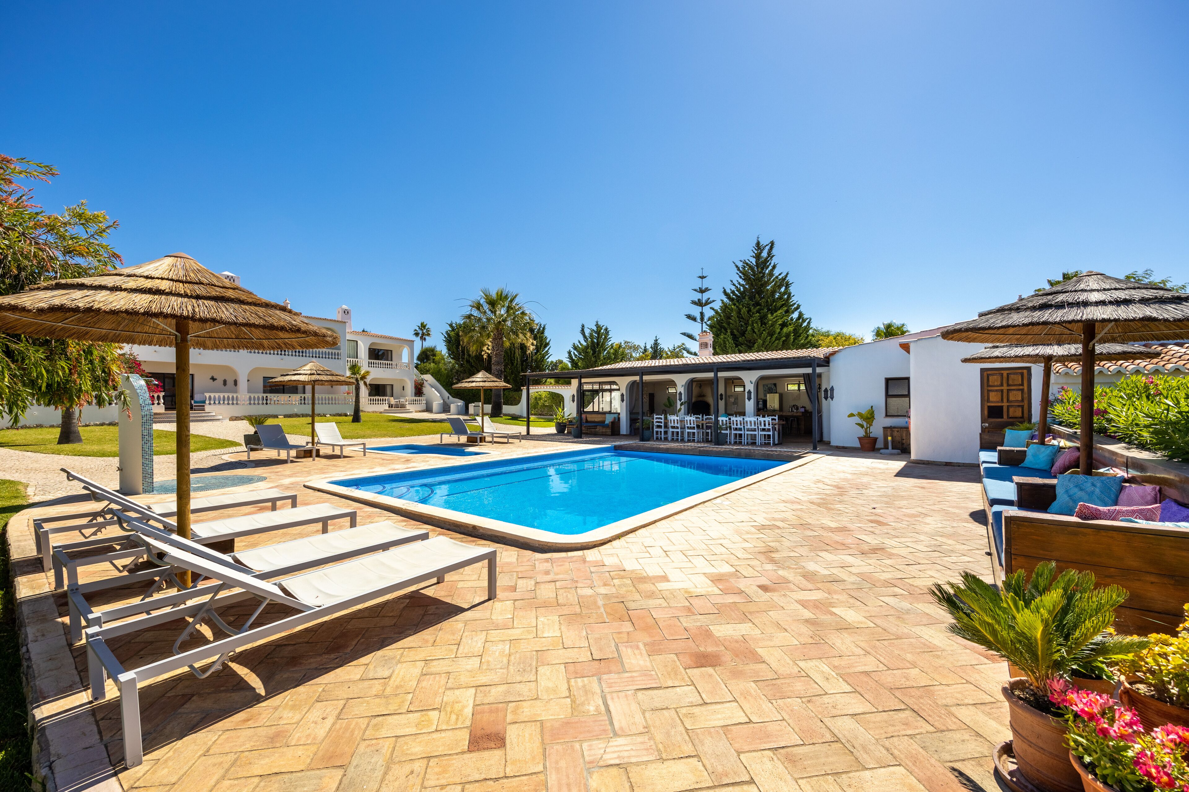 A pool area with lounge chairs and umbrellas, surrounded by a tiled patio and a building with a white facade.