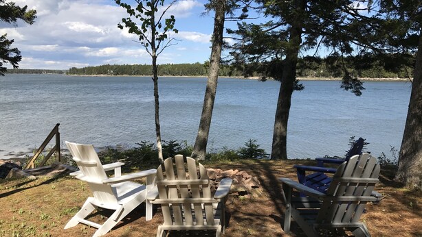 Une vue sur le lac, avec deux fauteuils Adirondack placés face à l’eau, entourés d’arbres et sous un ciel dégagé.