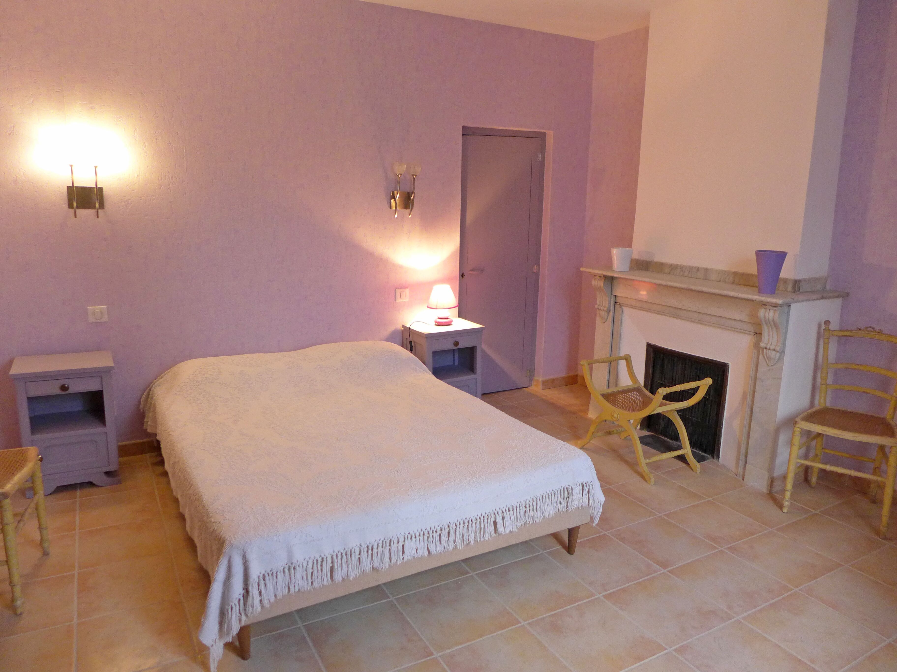 A bedroom with a bed, nightstands, a lamp, a chair, and a fireplace.