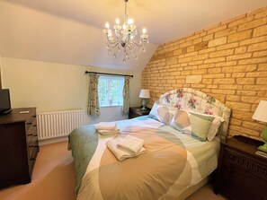 Un dormitorio con pared de ladrillo, una araña, cama con ropa de cama floral, un radiador y una ventana con cortinas.