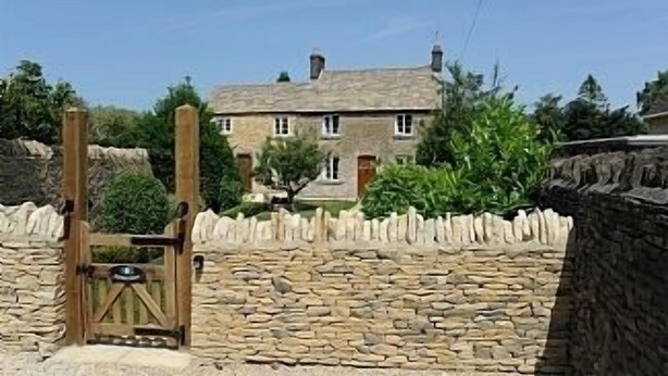 Un mur de pierre avec une porte en bois qui mène à un bâtiment en pierre doté de nombreuses fenêtres et d’un toit en pente.