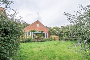 A brick house with a triangular roof, surrounded by a well-maintained lawn and dense foliage. - Betty's Cottage, Brancaster, Norfolk (Brancaster)