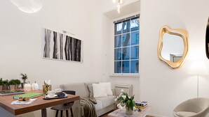 A modern living room with a sofa, coffee table, and a dining area with a table and chairs. There is a window with a view of a blue building outside. - Marvellous 3 bed / 2 bath apt near Vatican  (Roma)