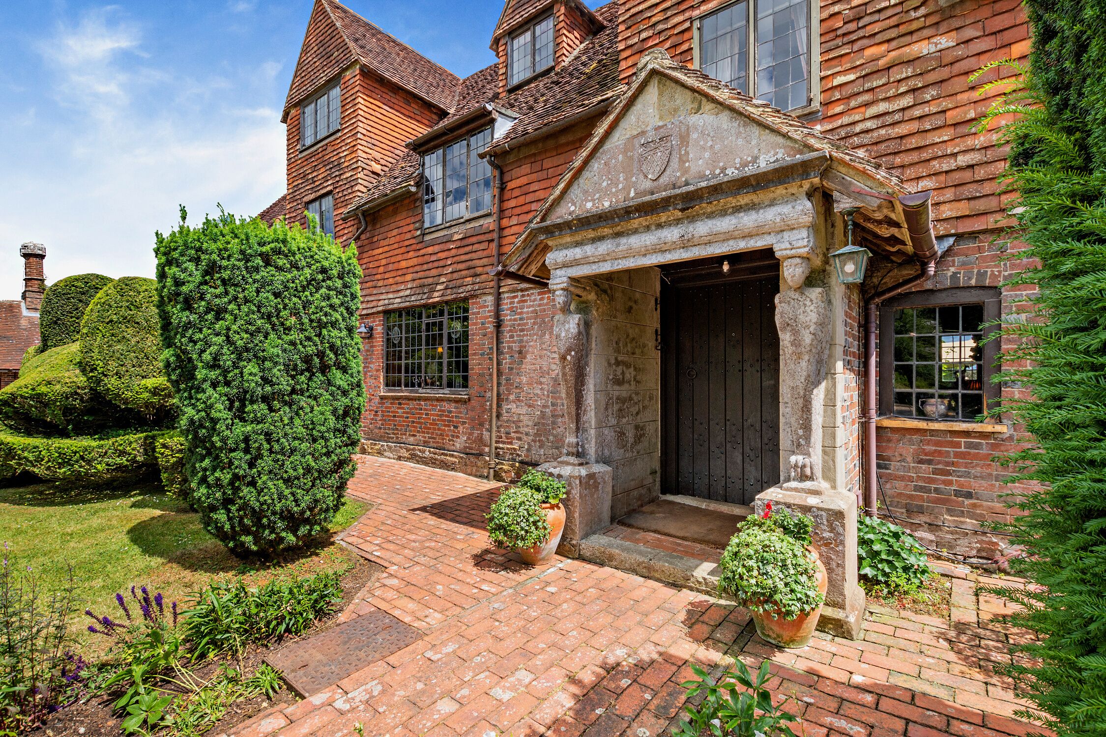 A traditional brick building with a symmetrical facade, a central entrance with a stone archway, and a well-maintained garden with hedges and potted plants.