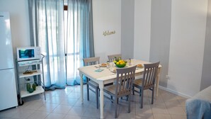 Un comedor con una mesa dispuesta para cuatro personas, un microondas, un estante pequeño con plantas y un cartel con la palabra 'Welcome' en la pared.