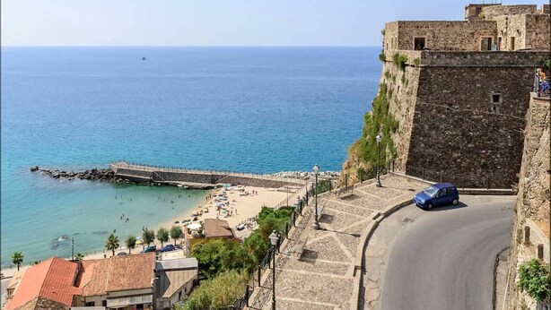Un pueblo costero con una fortificación de piedra, una playa con sombrillas y una vista despejada del mar.