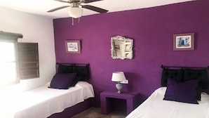 Una habitación con dos camas, un ventilador de techo, una mesita con lámpara y un espejo en la pared.
