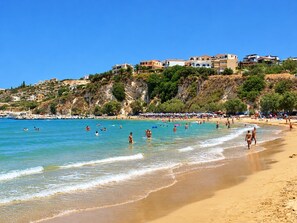 A beach with clear blue water, people swimming and walking on the sand, and buildings on a hillside in the background. - Private pool, & large garden - 100m from beach, tavernas & shops in Almyrida. (Almyrida)