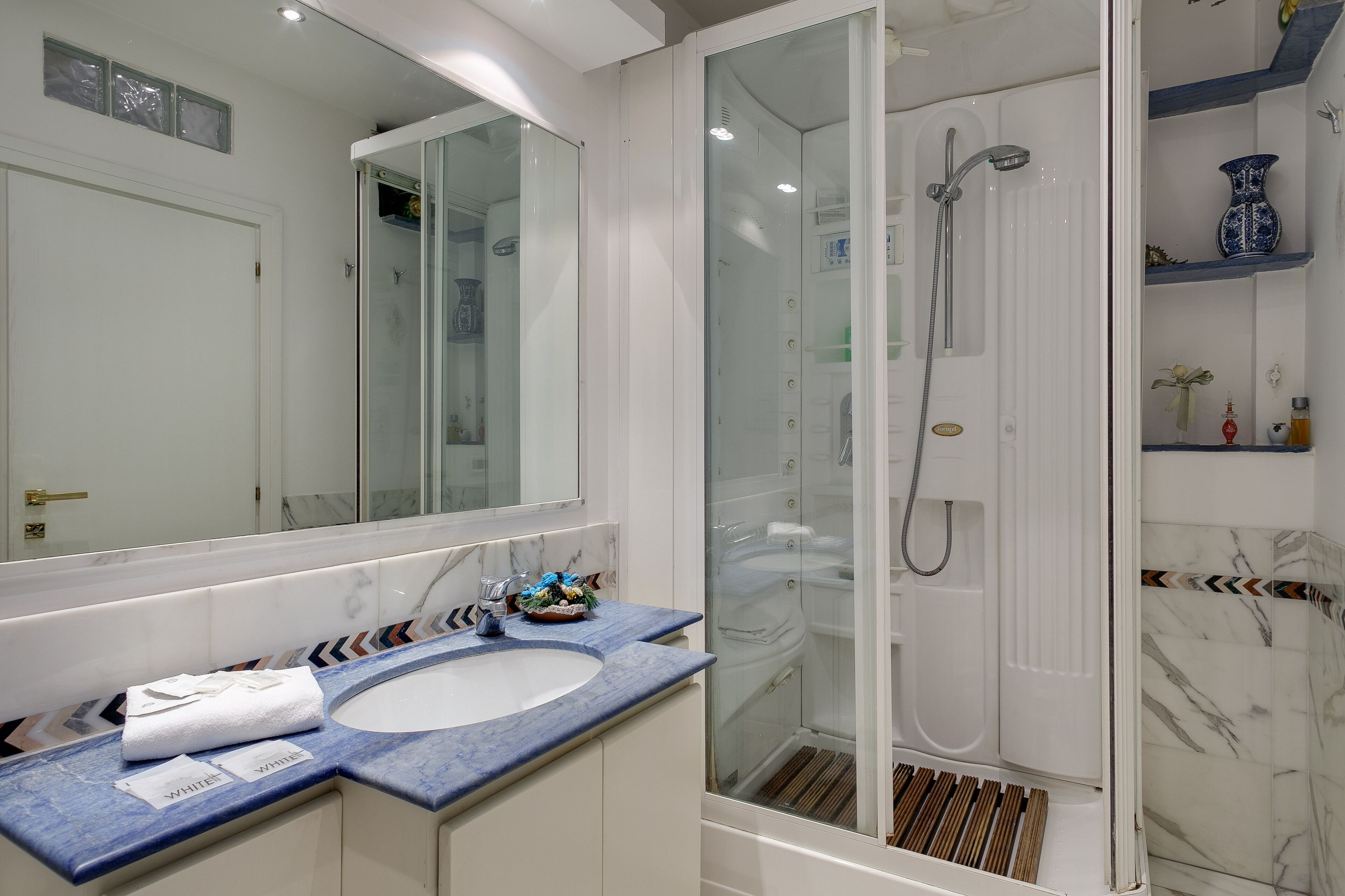 Une salle de bain avec un lavabo blanc, une douche à parois de verre et un mur recouvert de carreaux de marbre.