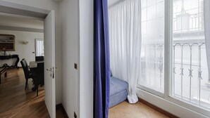 A room with a large window, a blue curtain, a bed, and a desk with a chair. - TREVI FOUNTAIN 3 BEDROOM APARTMENT PRESTIGIOUS. (Roma)