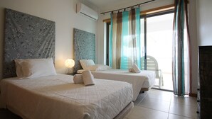 A hotel room with two beds, a wall-mounted air conditioner, and a view of the outside through a large window with curtains. - Varandas do Mar T1_T, frente mar, Olhos de Água (Albufeira)