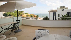 A terrace with a table and chairs, a large umbrella, and a view of the sea and buildings. - Varandas do Mar T1_T, frente mar, Olhos de Água (Albufeira)