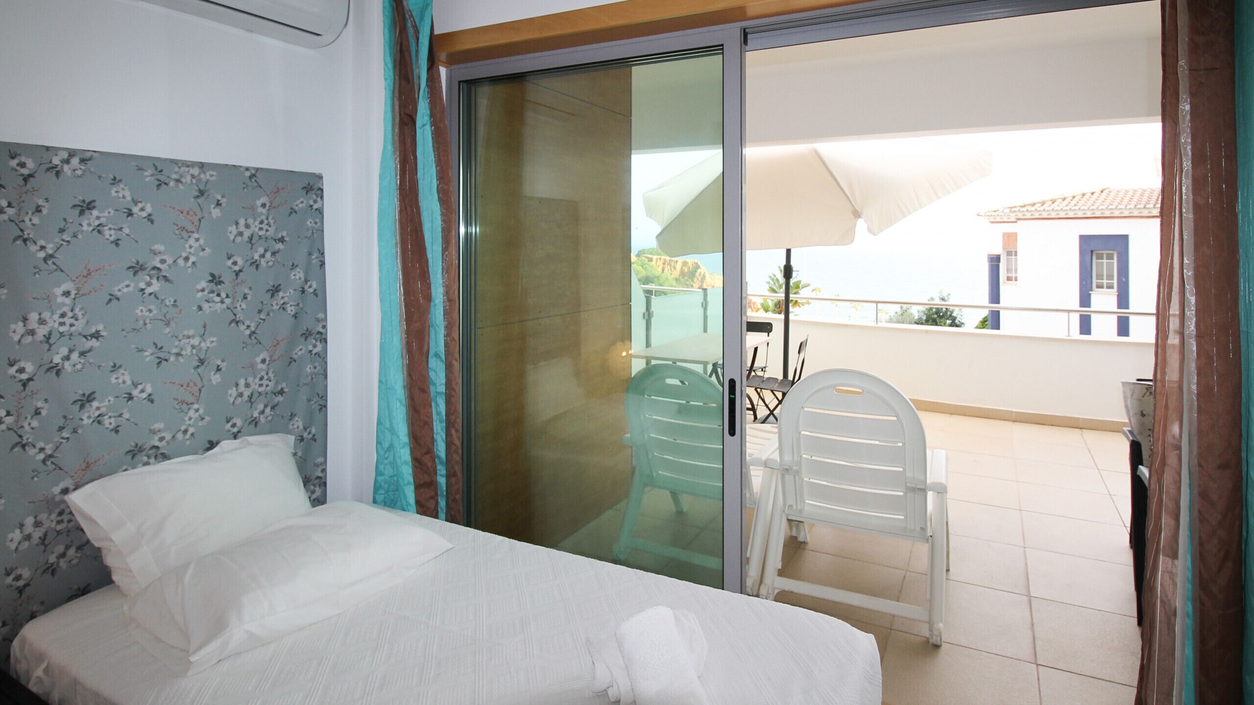 Une chambre avec un lit, une porte-fenêtre coulissante donnant sur un balcon équipé d’une table et de chaises, et une vue sur un immeuble situé de l’autre côté de la rue.