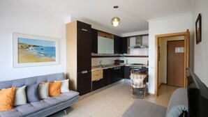 A modern kitchen with dark cabinetry, a dining area with a striped tablecloth, and a grey sofa with orange cushions. - Varandas do Mar T1_T, frente mar, Olhos de Água (Albufeira)