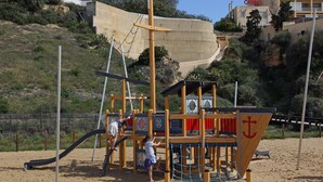 Une structure de jeu en bois sur le thème d’un navire, comprenant un mât et une ancre, située sur une plage de sable avec une clôture et des bâtiments en arrière-plan.
