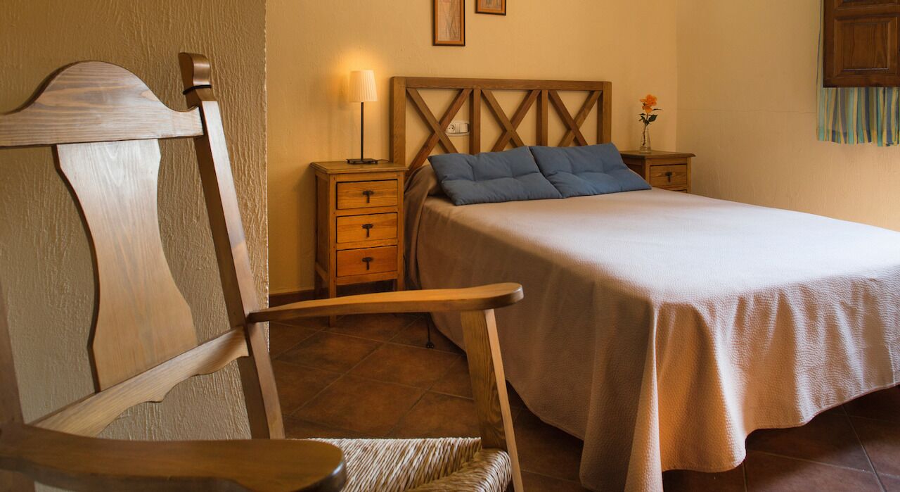 A bedroom with a wooden headboard, a bed with a beige bedspread and blue pillows, a nightstand with a lamp, and a small table with a vase of flowers.