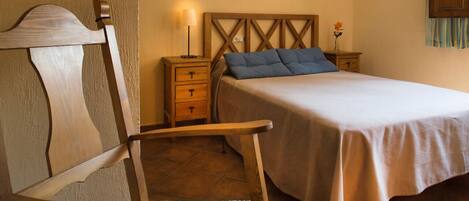 A bedroom with a wooden headboard, a bed with a beige bedspread and blue pillows, a nightstand with a lamp, and a small table with a vase of flowers.