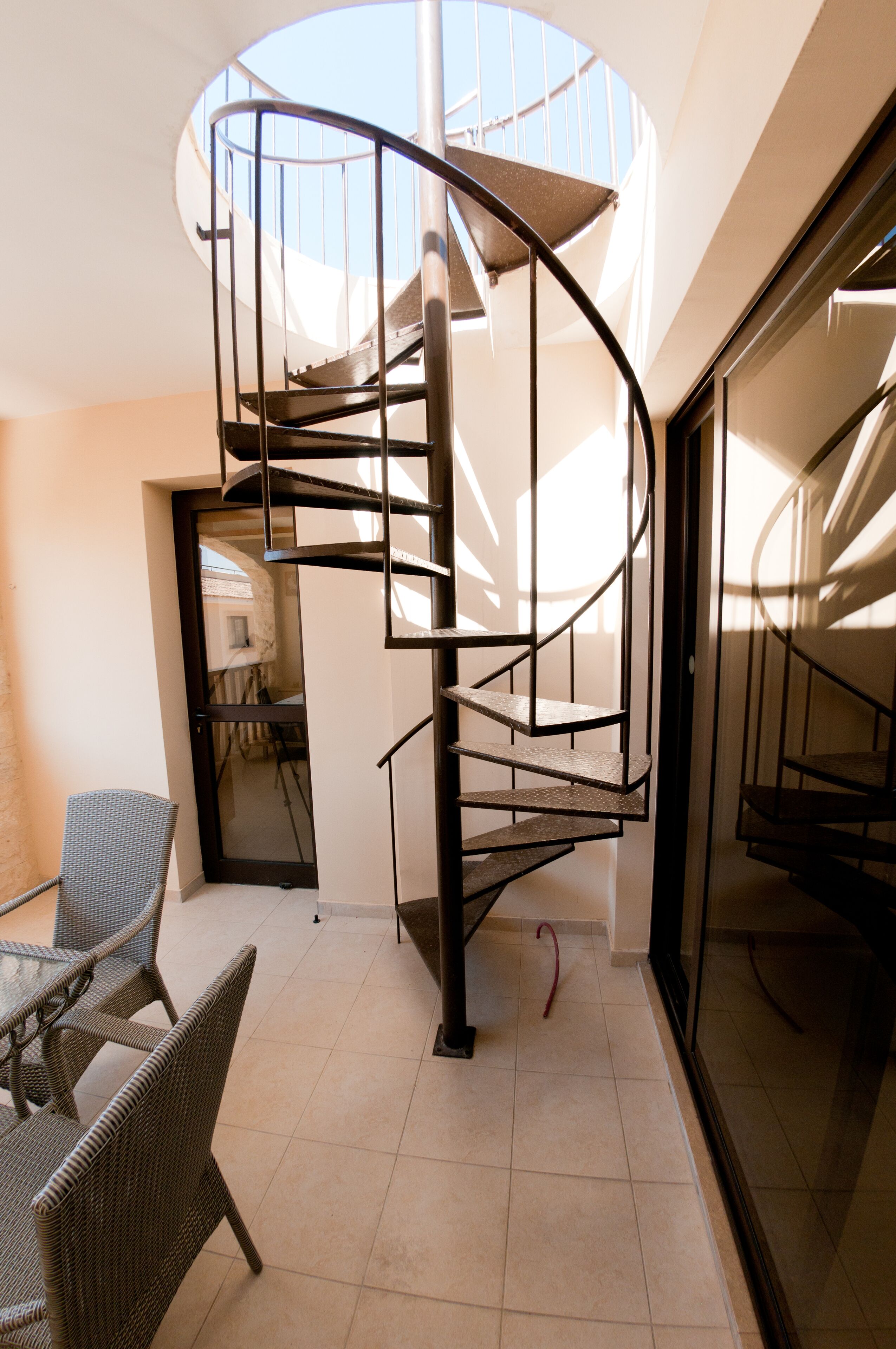 Una escalera en espiral en un interior moderno con mobiliario de mimbre y un ventanal que da a otra habitación.