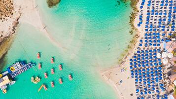 Vista aérea de una playa con aguas turquesas cristalinas, un muelle con barcos y filas de sombrillas azules.