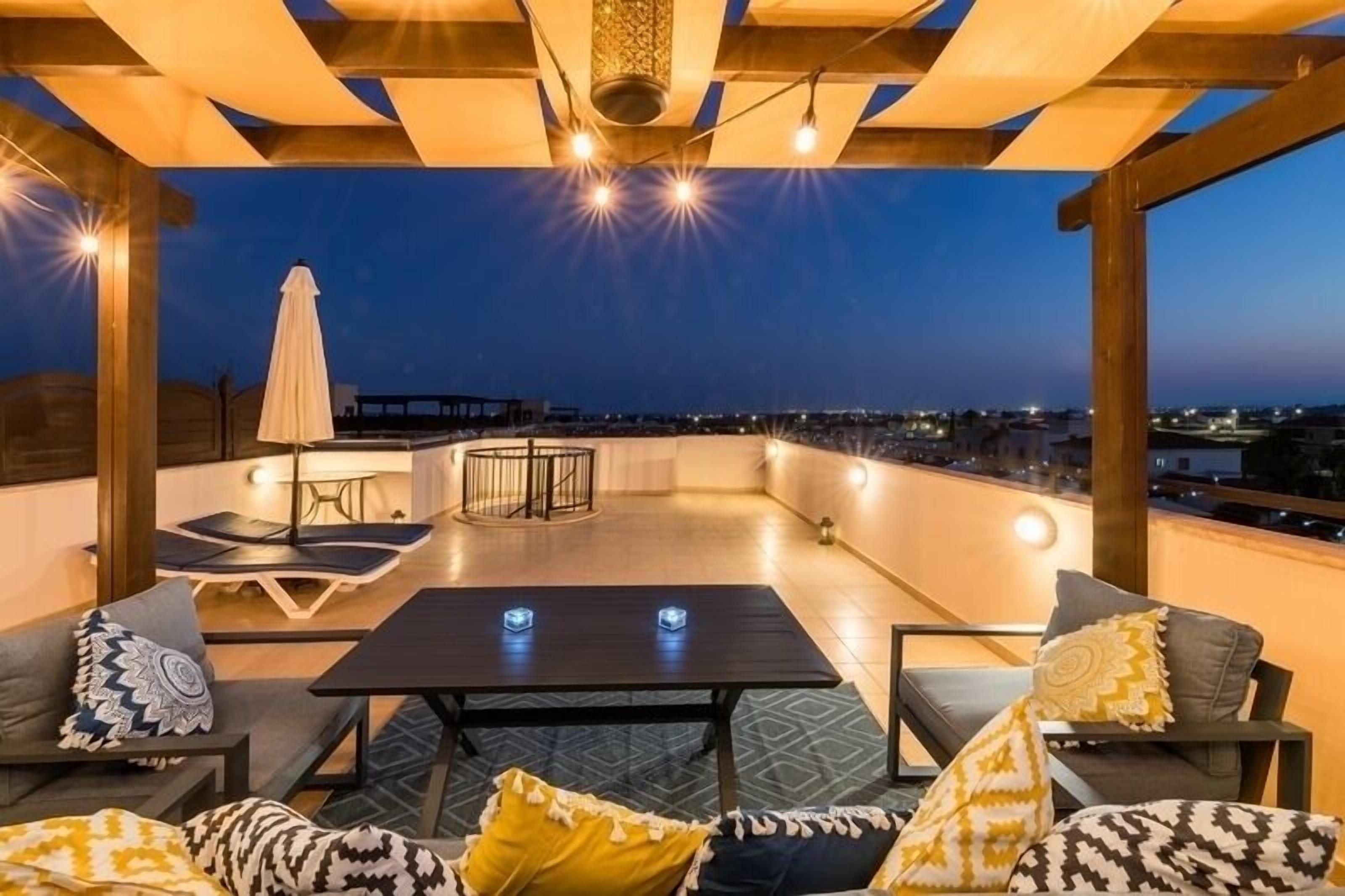 Una terraza en la azotea con vistas a la ciudad al anochecer, que incluye una mesa, sillas y un sofá estampado.