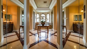 A spacious hallway with a central round table, a large mirror, and a view of a kitchen with a window.