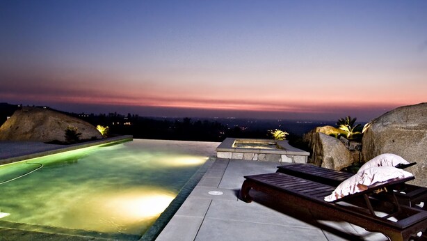 A swimming pool with a lounge chair and a stone bench, surrounded by rocks and a sunset view.