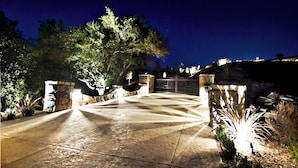 A paved driveway leading to a gated entrance with stone walls and a large tree.