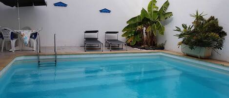 A swimming pool with lounge chairs and umbrellas, surrounded by plants and a white wall.