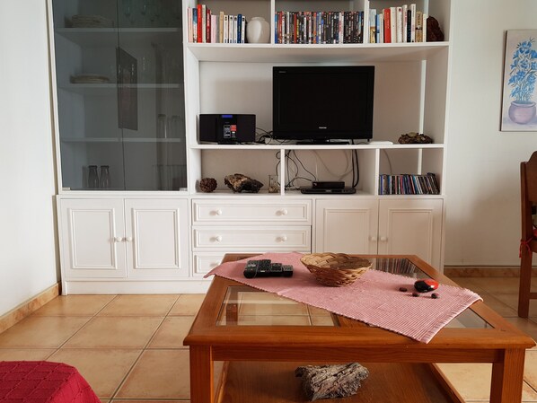 A living room with a glass-top coffee table, a bookshelf with books and DVDs, a flat-screen TV, and a decorative vase. - Nice house, private pool, WIFI, near to the beach. (Caleta de Famara)